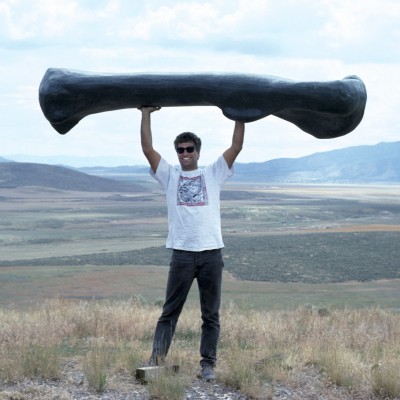 Louie Psihoyos getting a work out with a cast of a giant sauropod bone while wearing a Ray Troll t-shirt. &copy; Louis Pishoyos 1994
&nbsp;