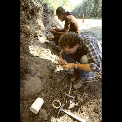 A younger Hans Sues sitting on a bucket happily digging fossils in a roadside cut.
